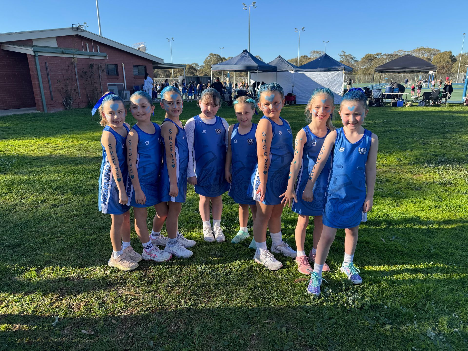 A netball team of young girls in blue dresses stands on a grass field in front of a building.