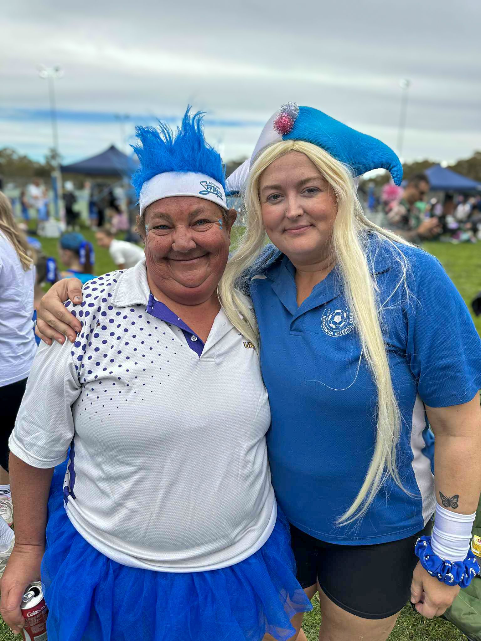 Two women in blue and white sports attire pose outdoors, smiling. One has blue spiky hair, the other a blue hat and long blonde wig.