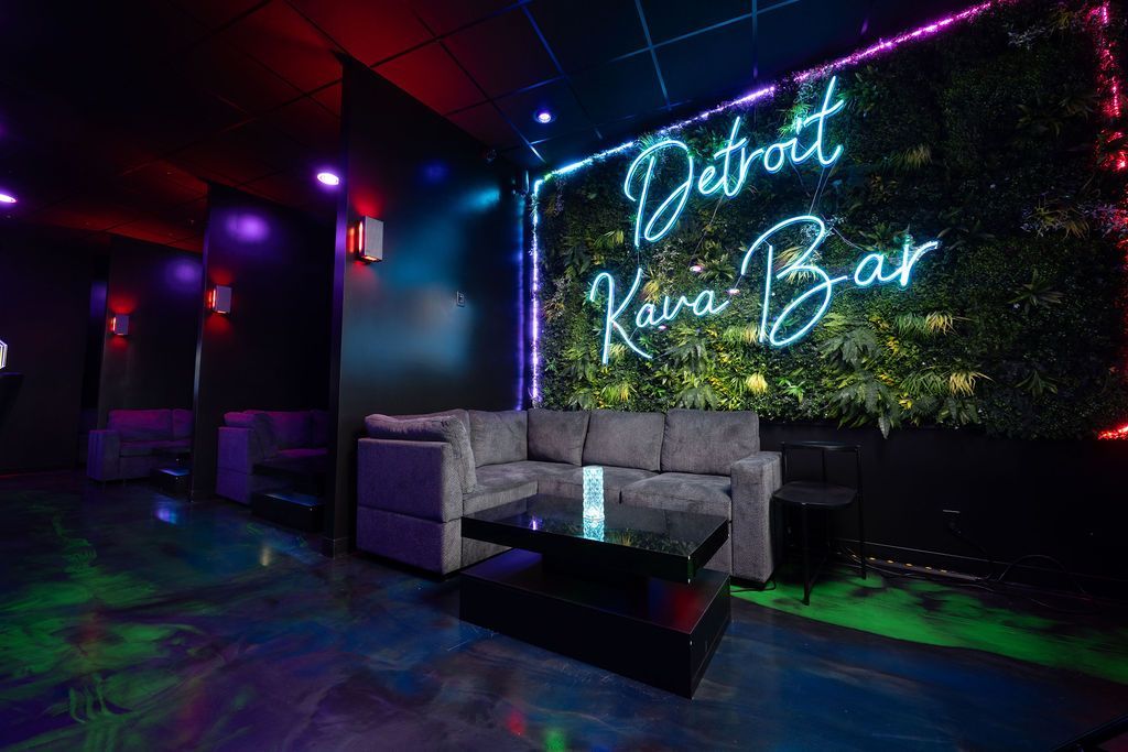 Neon-lit lounge with gray sofas and a glowing “Detroit Kava Bar” sign on a plant wall.