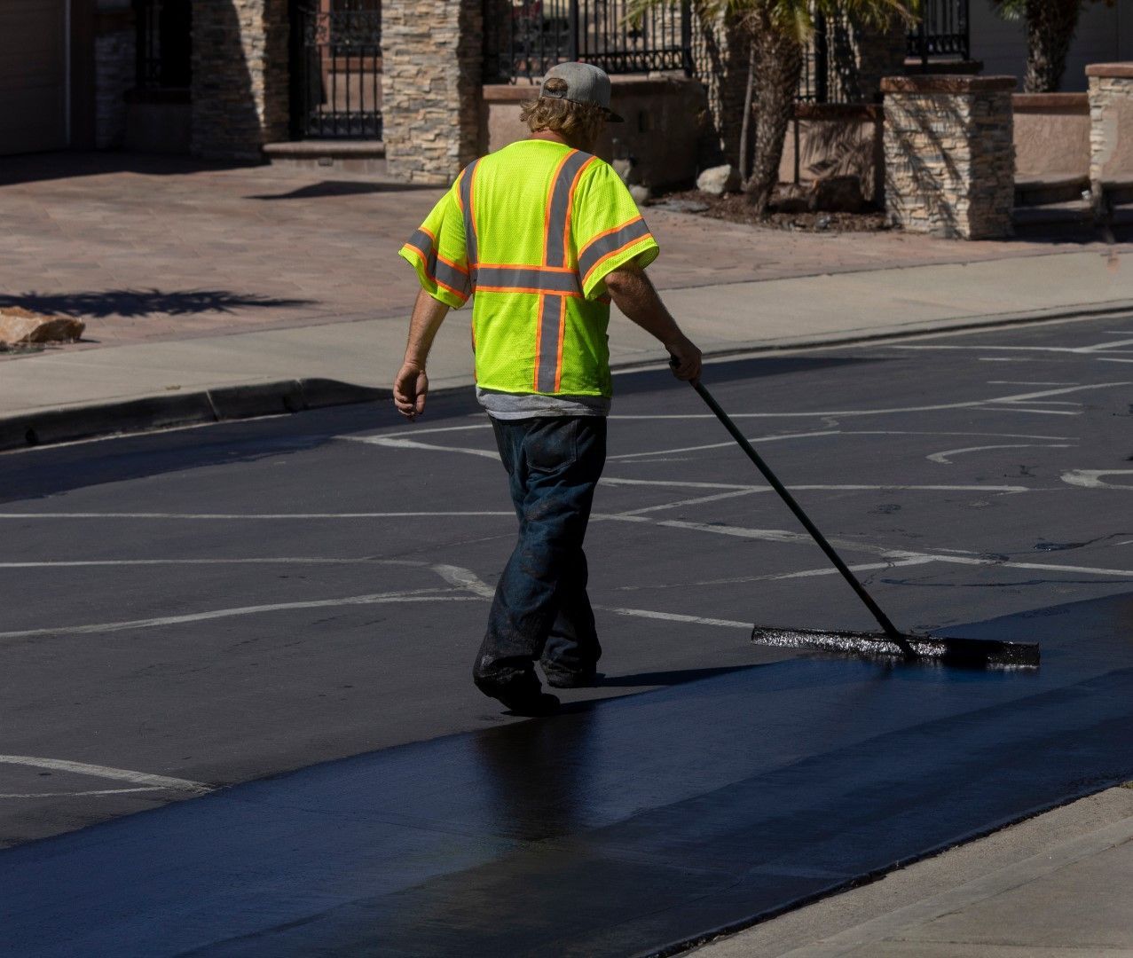 A person in a reflective vest applies sealant to a paved surface. The person is holding a long-handled tool in an outdoor setting.