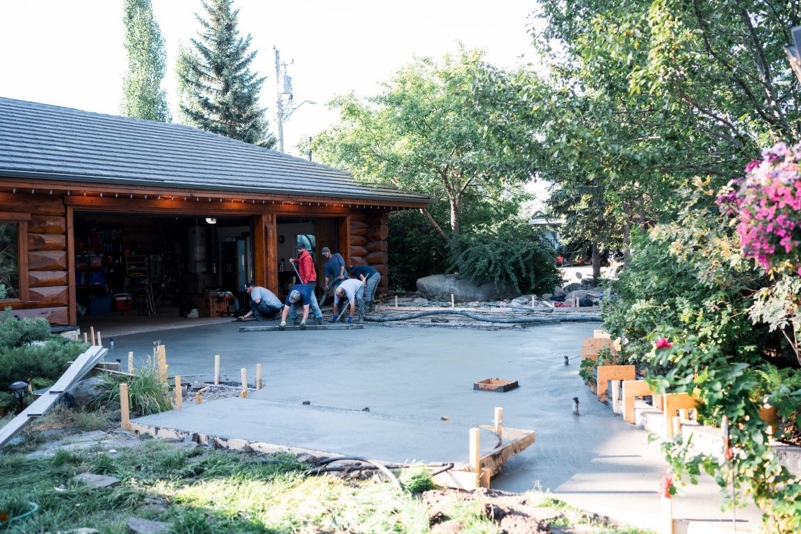 Workers pouring concrete for a driveway in front of a wooden garage. Freshly poured concrete is visible, with wooden framing and lush greenery in the background.