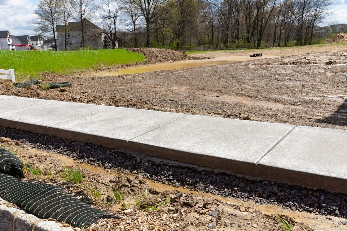 Newly poured concrete sidewalk beside muddy construction site with green grass in the background.