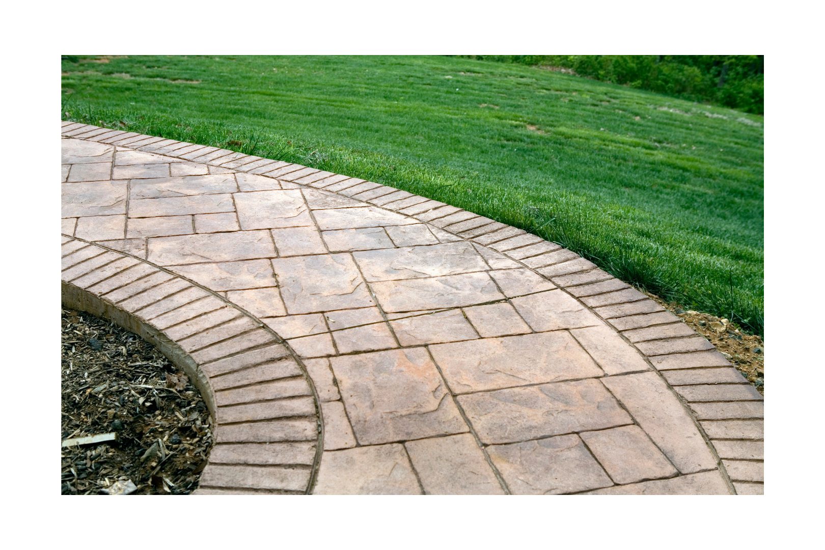 Curved paved driveway lined with green shrubs and bark mulch, adjacent to a concrete wall.