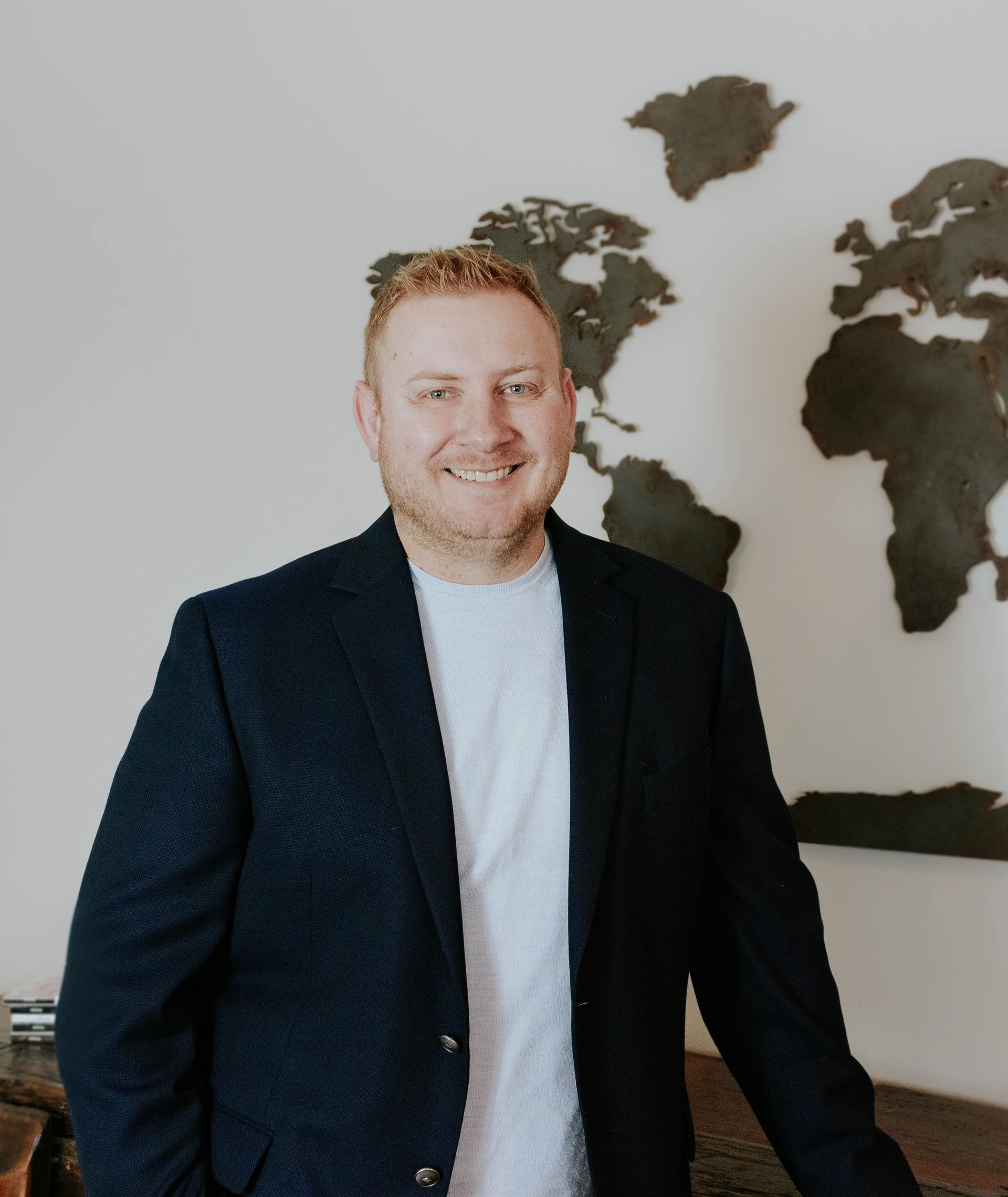 Man in navy blazer and white shirt smiles, standing in front of a world map.