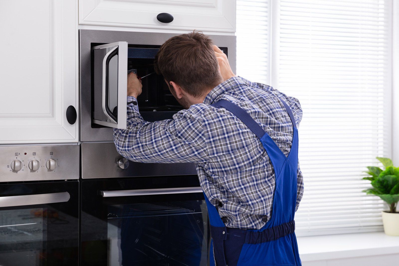 A person in blue overalls repairs a microwave oven in a kitchen.