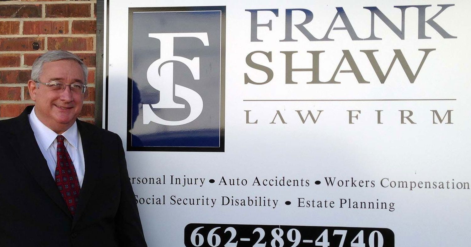 Lawyer Frank Shaw standing in front of his law firm's sign. The sign lists services and a phone number.