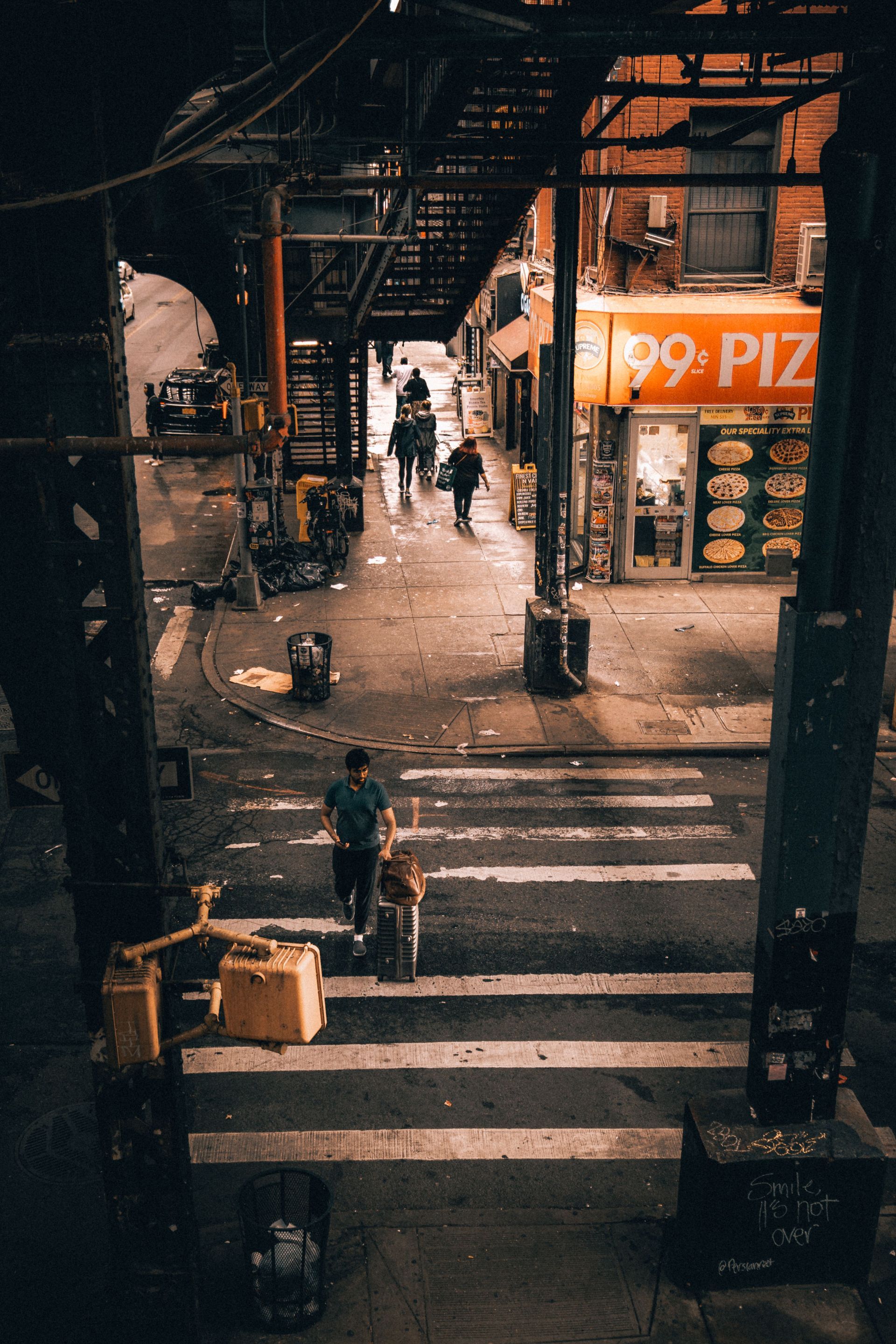 Rainy city crosswalk under an elevated train, with a cyclist and pedestrians near a lit storefront.