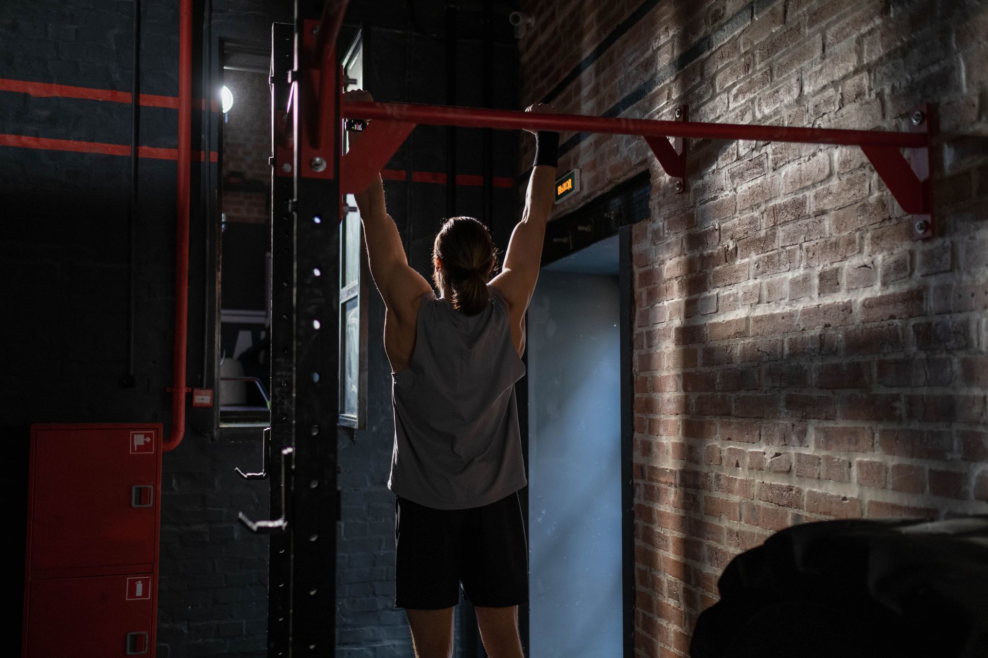 Person hanging from a pull-up bar in a dim gym with brick walls and red equipment