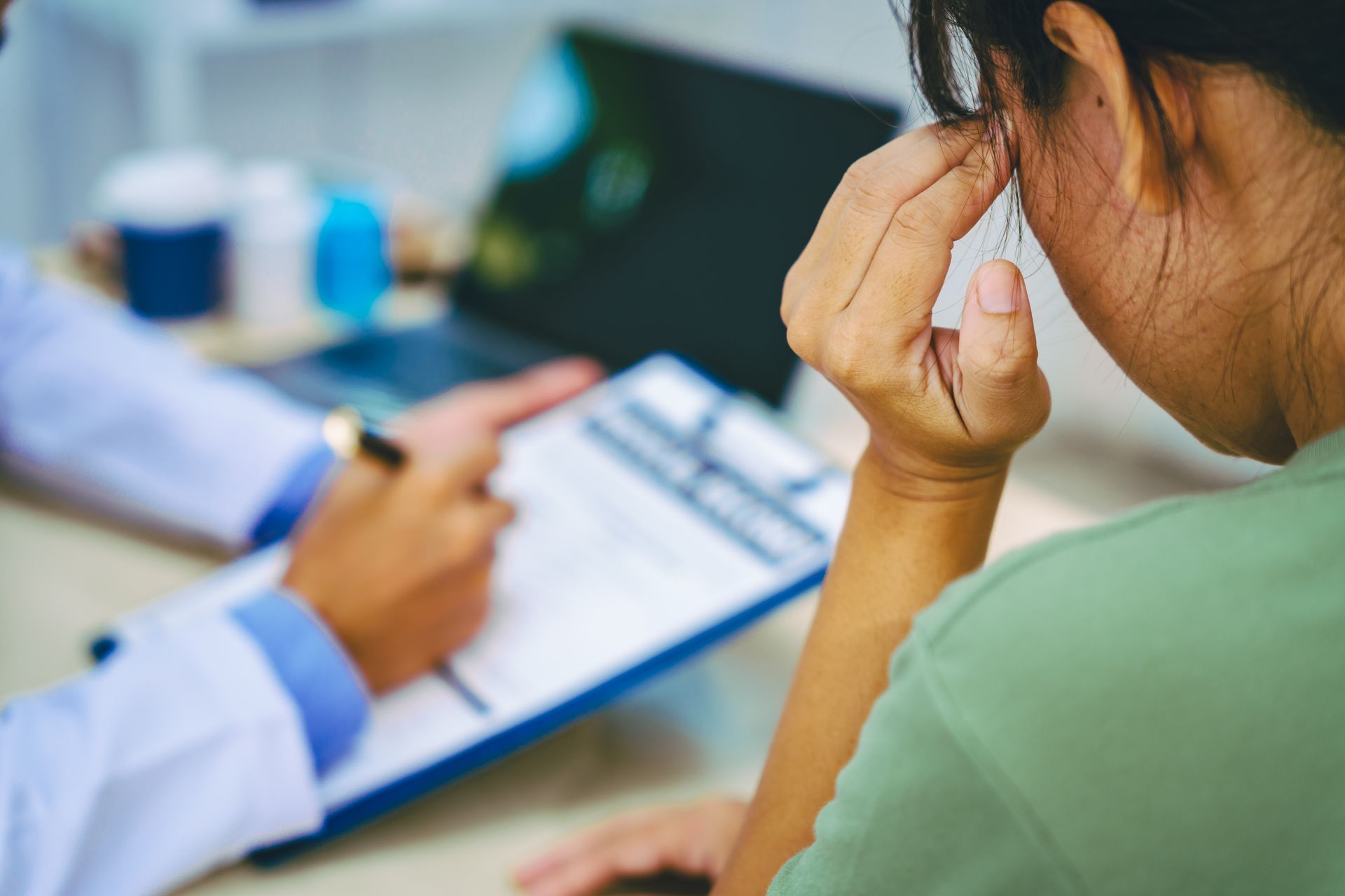 A person sits with their hand on their forehead while a doctor writes on a clipboard during a consultation.