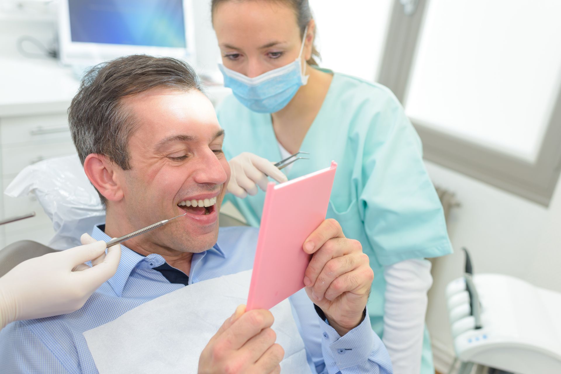 A dental professional in a mask and gloves holds a tool as a patient smiles while viewing his teeth in a pink mirror.