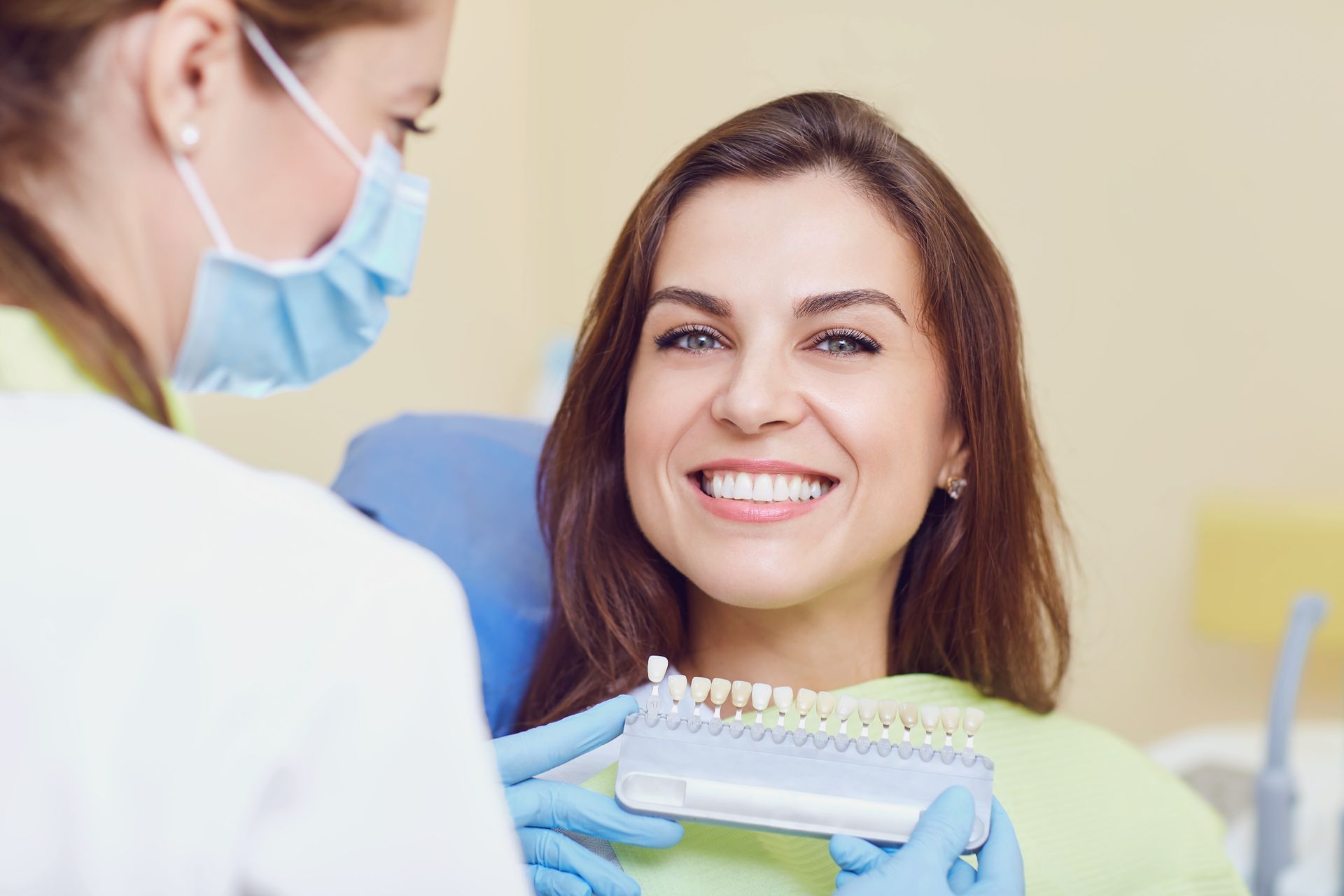 A dentist in a mask and gloves holds a tooth shade guide for a patient smiling in a dental chair.