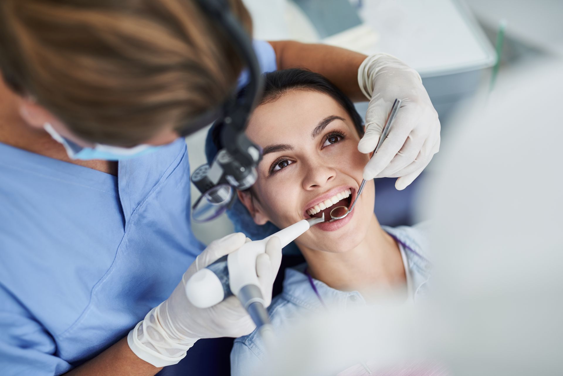 A dentist wearing a mask, face shield, and blue gloves examines a patient in a dental chair under a surgical light.