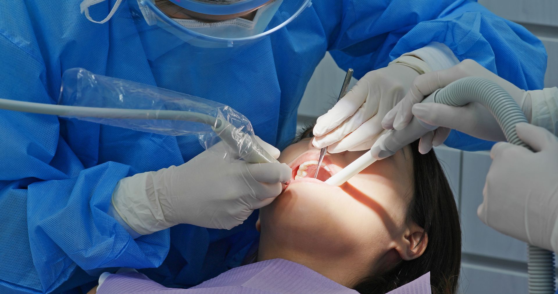 A dentist in blue scrubs and a face shield performs a procedure on a patient in a dental chair.