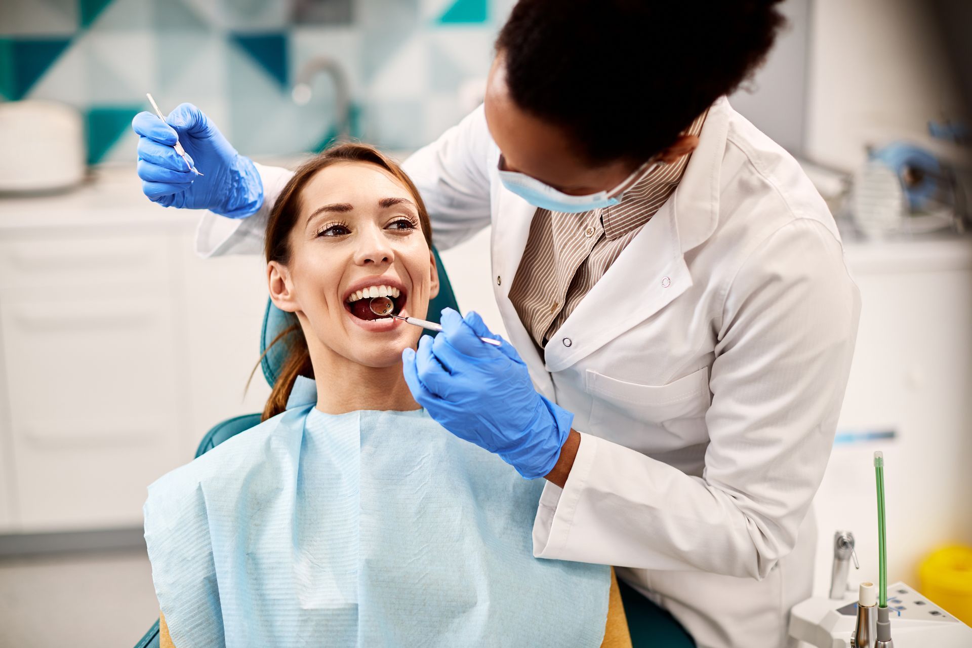 A dentist wearing blue gloves and a mask examines the teeth of a smiling patient in a dental chair.
