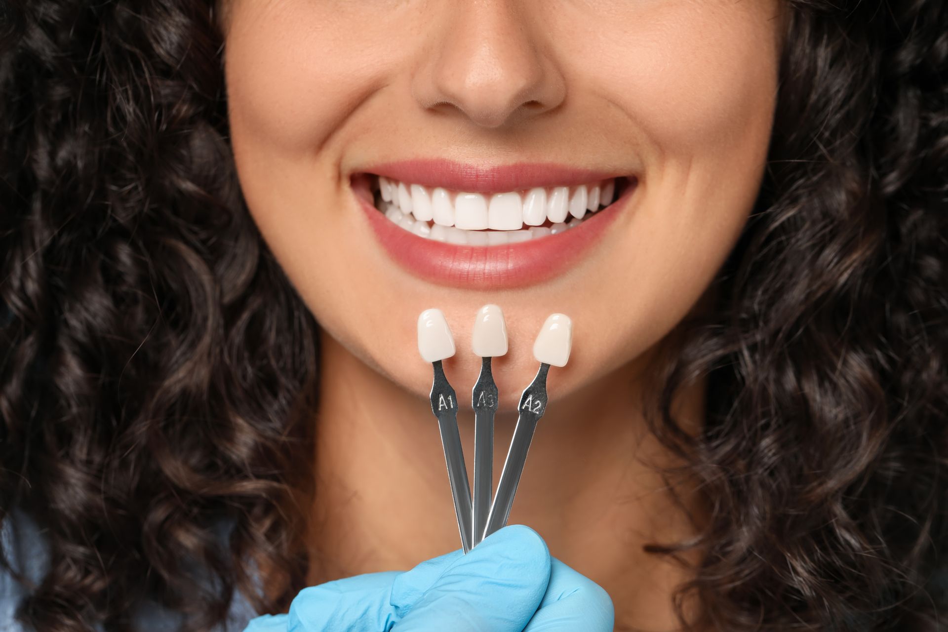 A person’s smile is being compared against three porcelain tooth shade tabs held by a gloved hand.