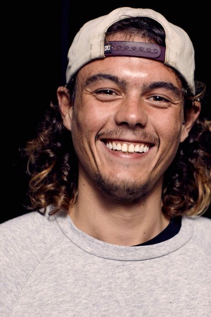 A man with long curly hair is wearing a hat and smiling.