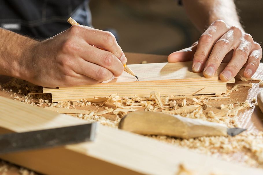 Hands carving a wooden board with a chisel, surrounded by wood shavings on a workbench