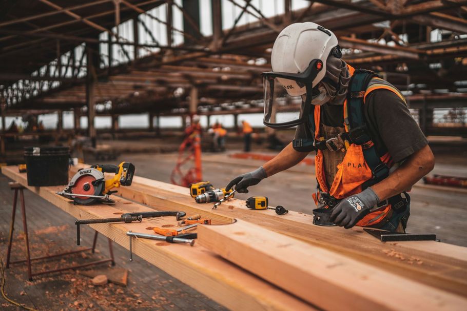Worker in a hard hat measuring lumber in a construction site workshop with power tools on a bench