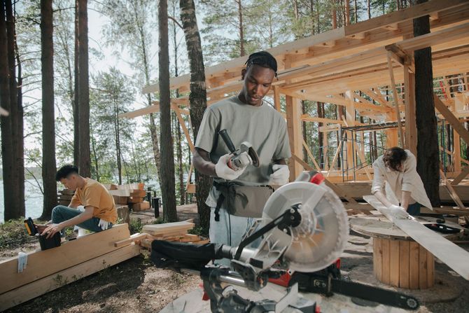 Workers building a wooden structure in a forest, with a man cutting boards using a miter saw.