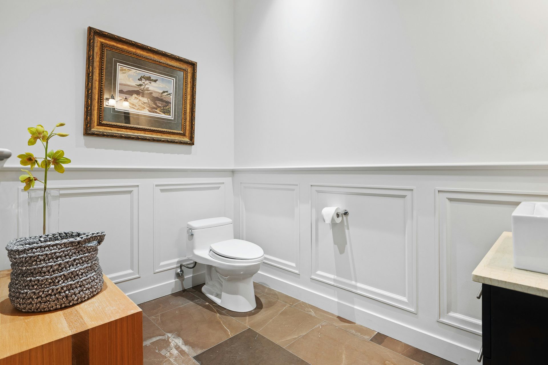 Small bathroom with white wainscoting, toilet, wall art, and a wicker basket on a wooden table.