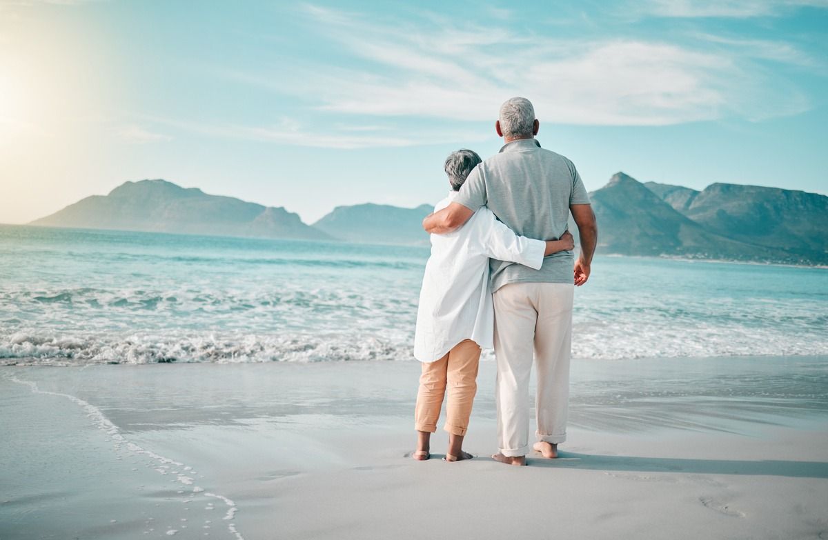 Couple standing arm-in-arm on a sandy beach, looking out at calm ocean and distant mountains.