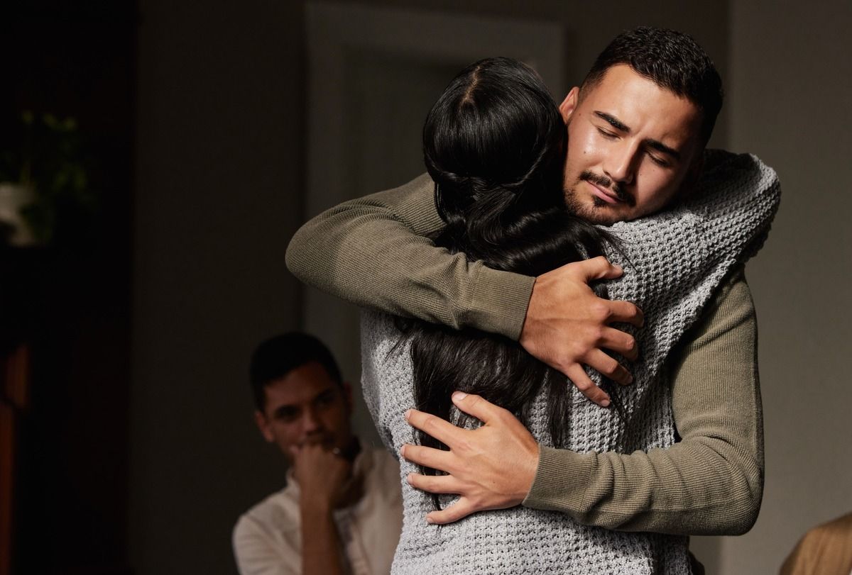 Two people embrace tightly in a dim room while a third person stands in the background.