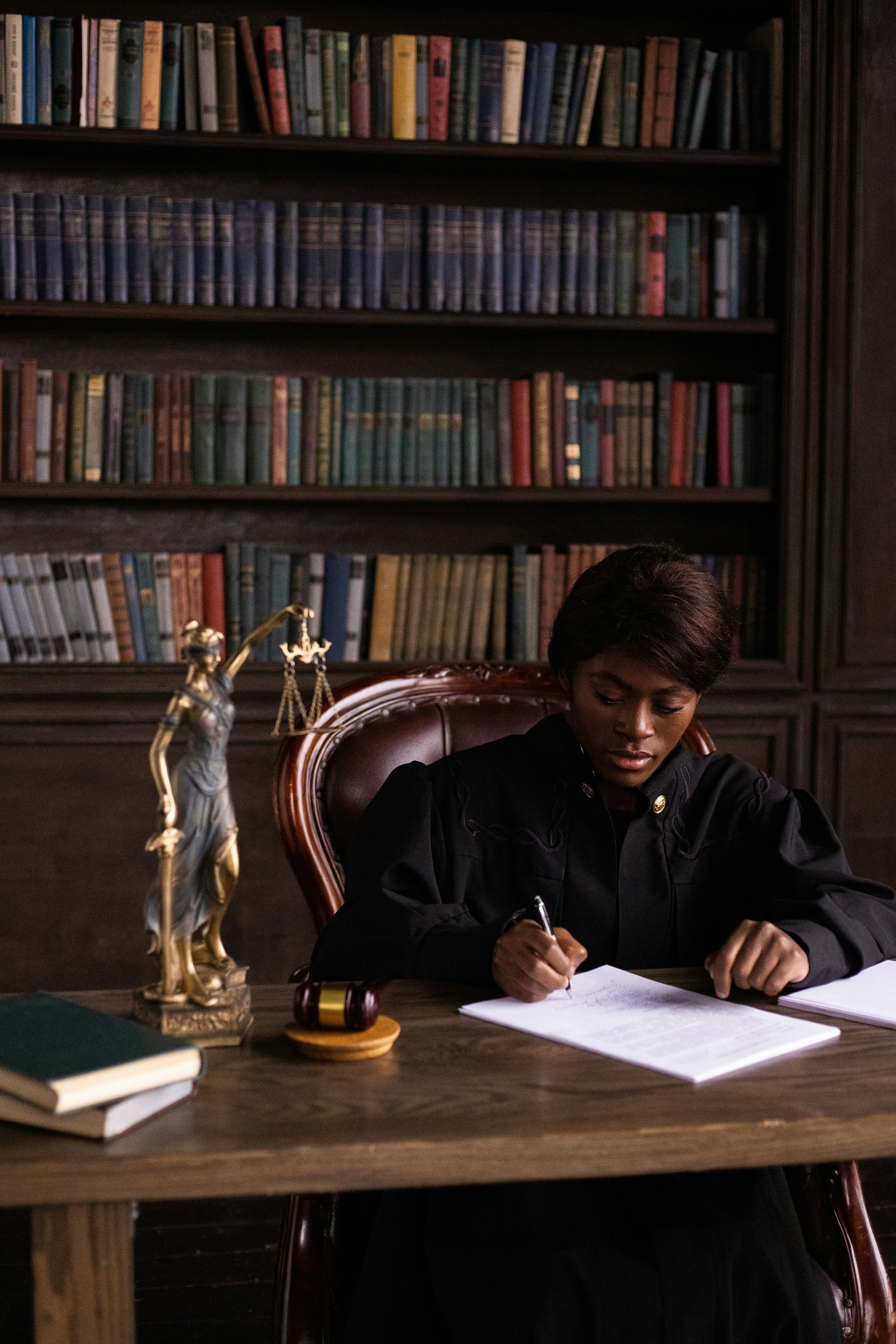 Judge in black robe writing at a desk with scales of justice statue and library background.