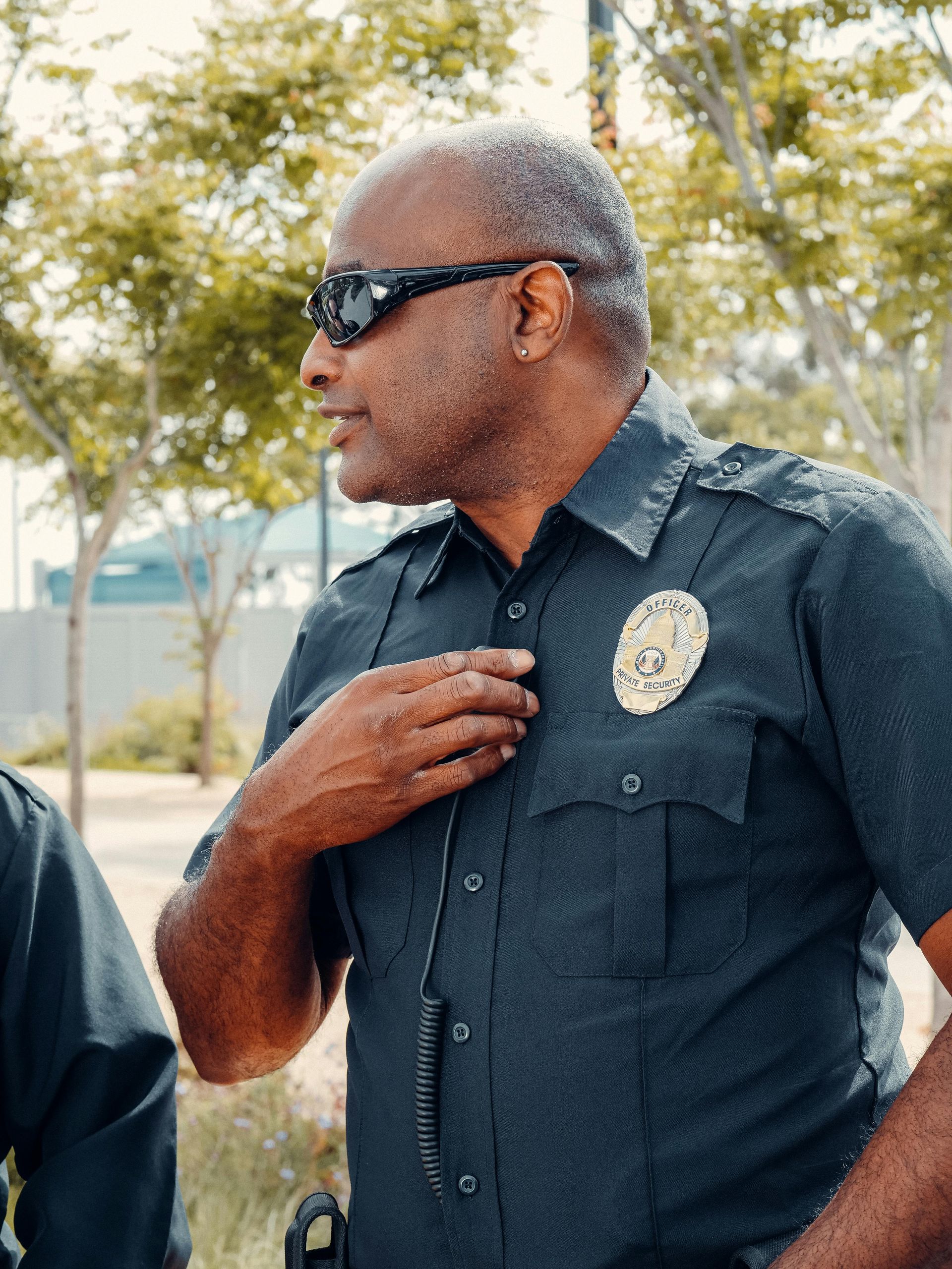 Police officer wearing sunglasses, adjusting his shirt near a park.