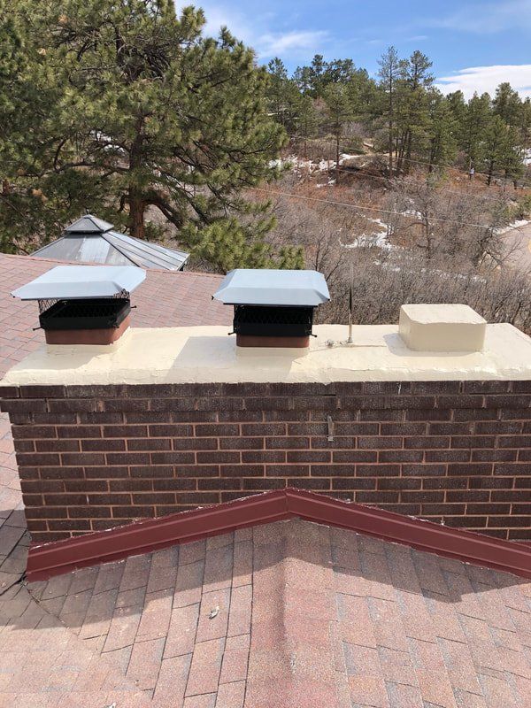 Brick chimney with two metal chimney caps on a roof, with trees in the background.