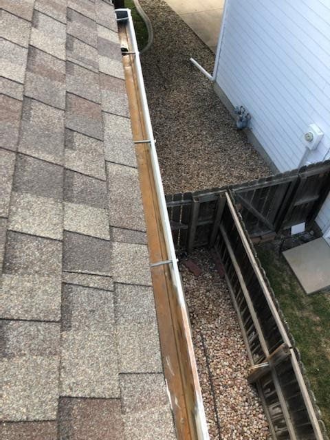 View of roof shingles, gutter, and gravel path alongside a building and wooden fence.