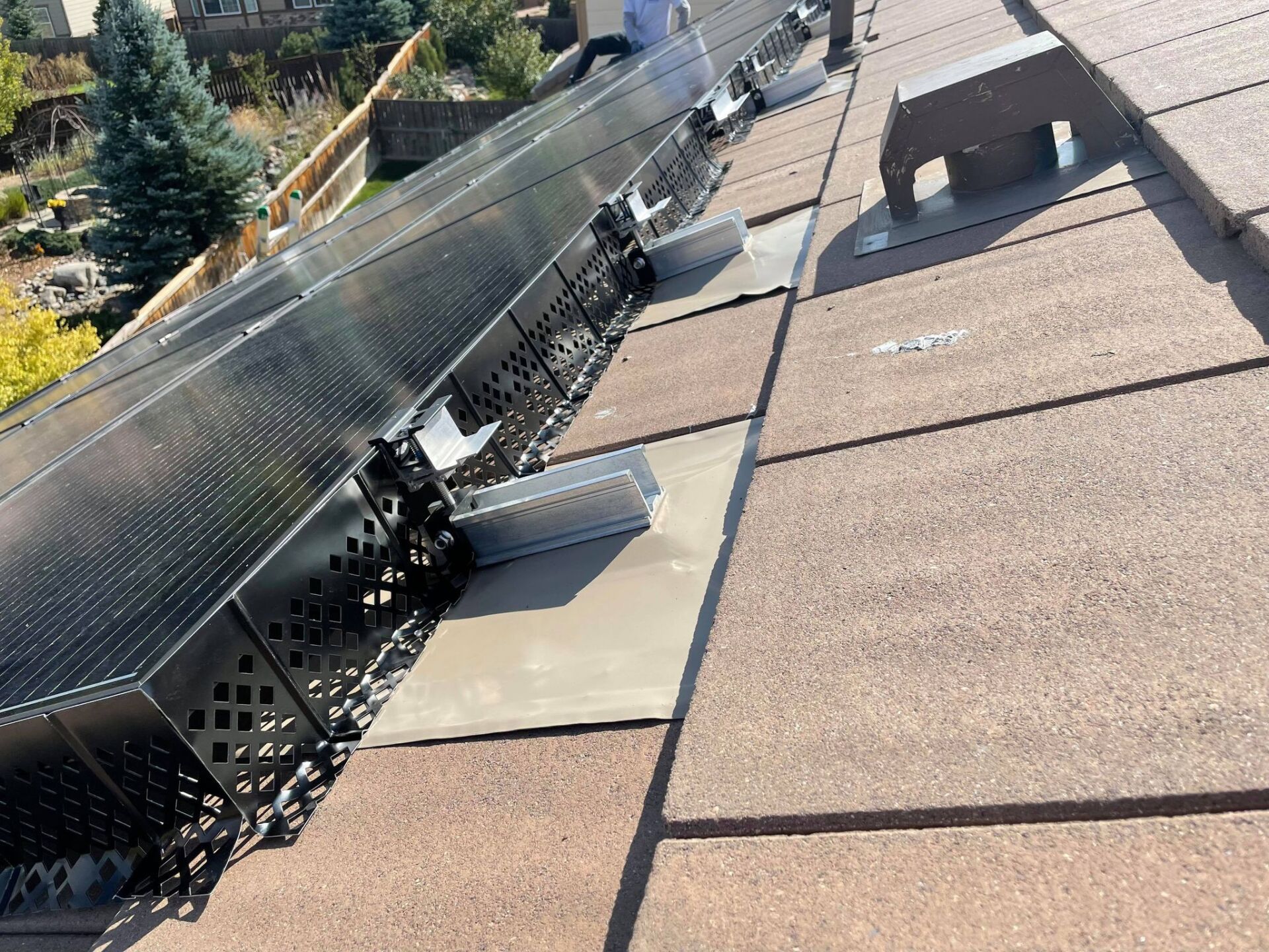 Solar panels installed on a brown tiled roof. Panels are dark, attached to metal mounting hardware. Sunny outdoor setting.