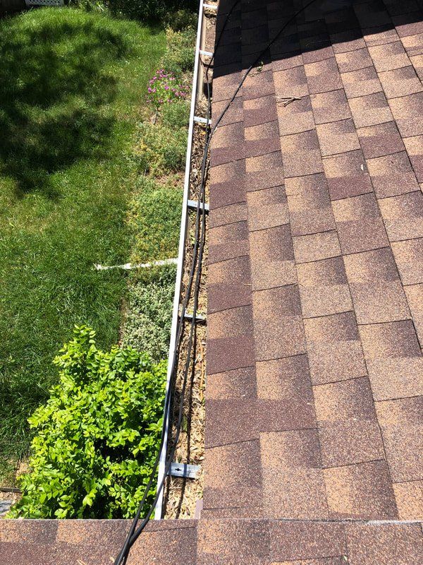 Brown shingled roof with a gutter and cables running along it, overlooking green lawn and a bush.