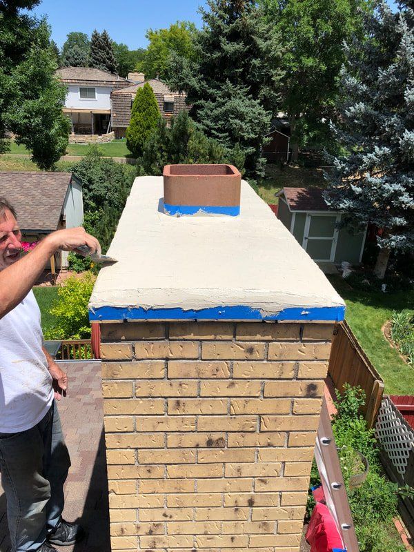 Man applying sealant to a chimney. White sealant over blue tape. Brick chimney with a terracotta flue cap.