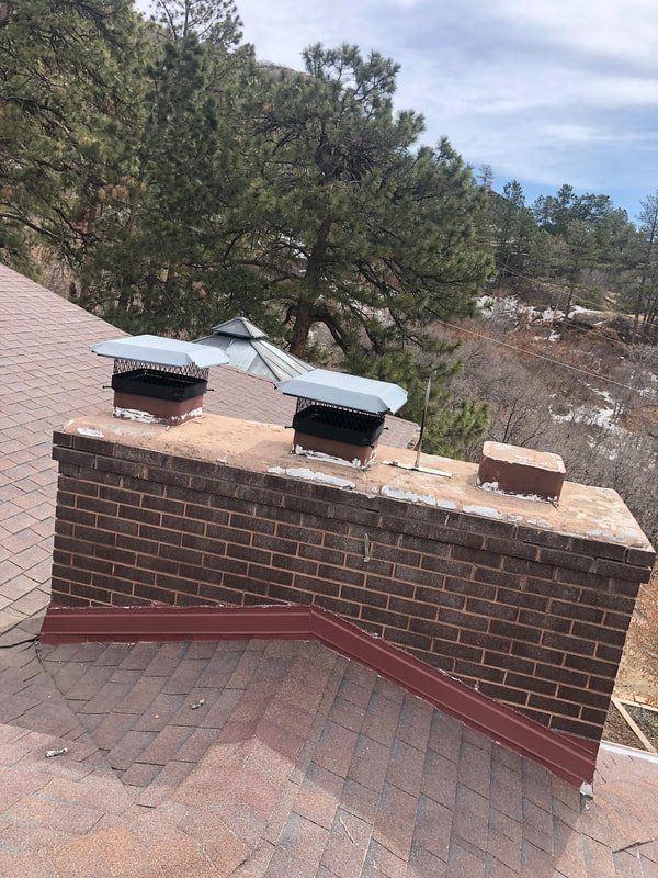 Brick chimney with two metal chimney caps on a shingled roof, surrounded by trees.