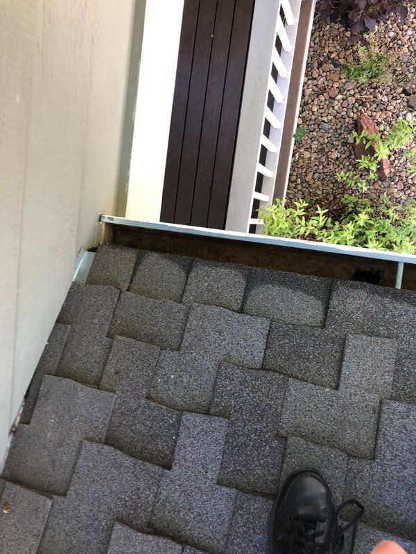 Overhead view of a roof with gray shingles. A gutter runs along the edge, adjacent to a beige wall and dark wooden trim.