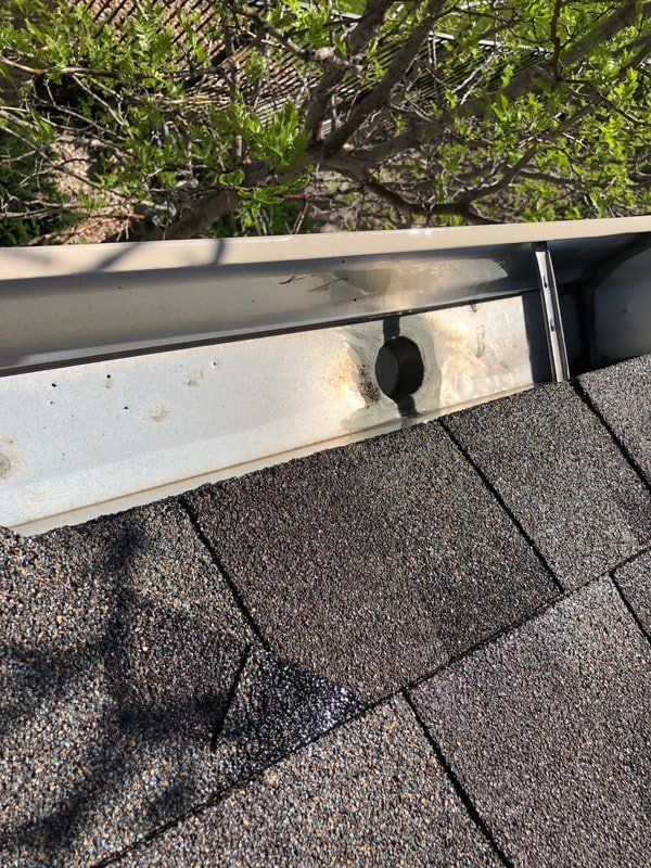 A close-up of a roof and gutter. The gutter is light gray, with a drain hole.  Shingles are brown and black.