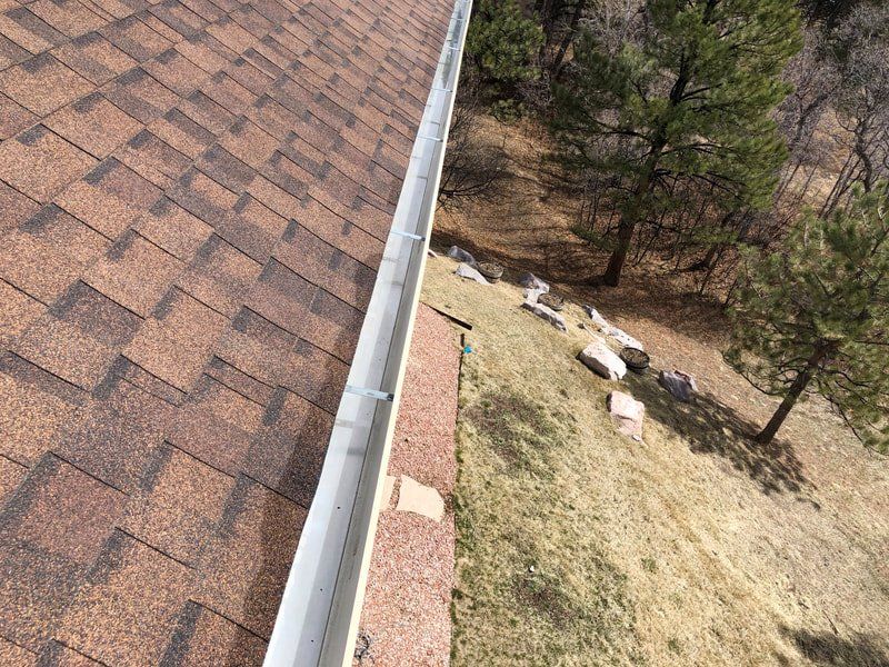 Brown shingled roof with a white gutter, adjacent to a grassy hillside and trees.