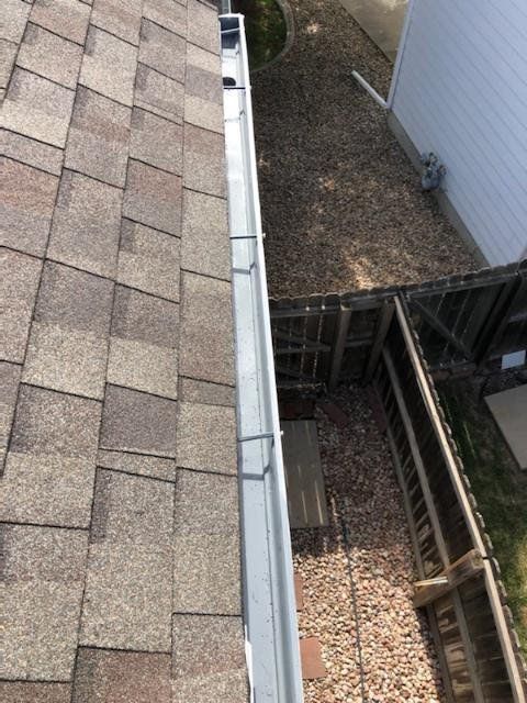 Close-up of a house roof with brown shingles, a silver gutter, and a gravel-covered yard below.