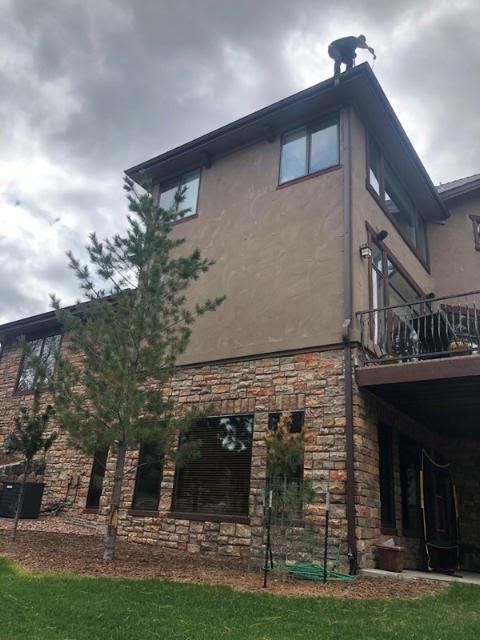 Person on a roof cleaning gutters of a two-story house with stone and stucco siding, brown trim, and a cloudy sky.