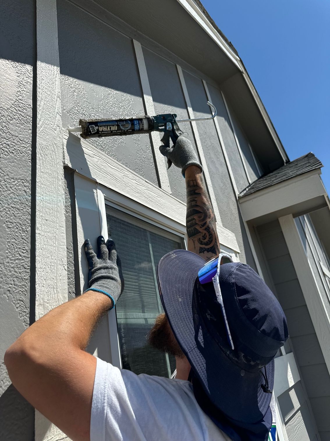 A man wearing a hat and gloves is applying caulk to the side of a building.