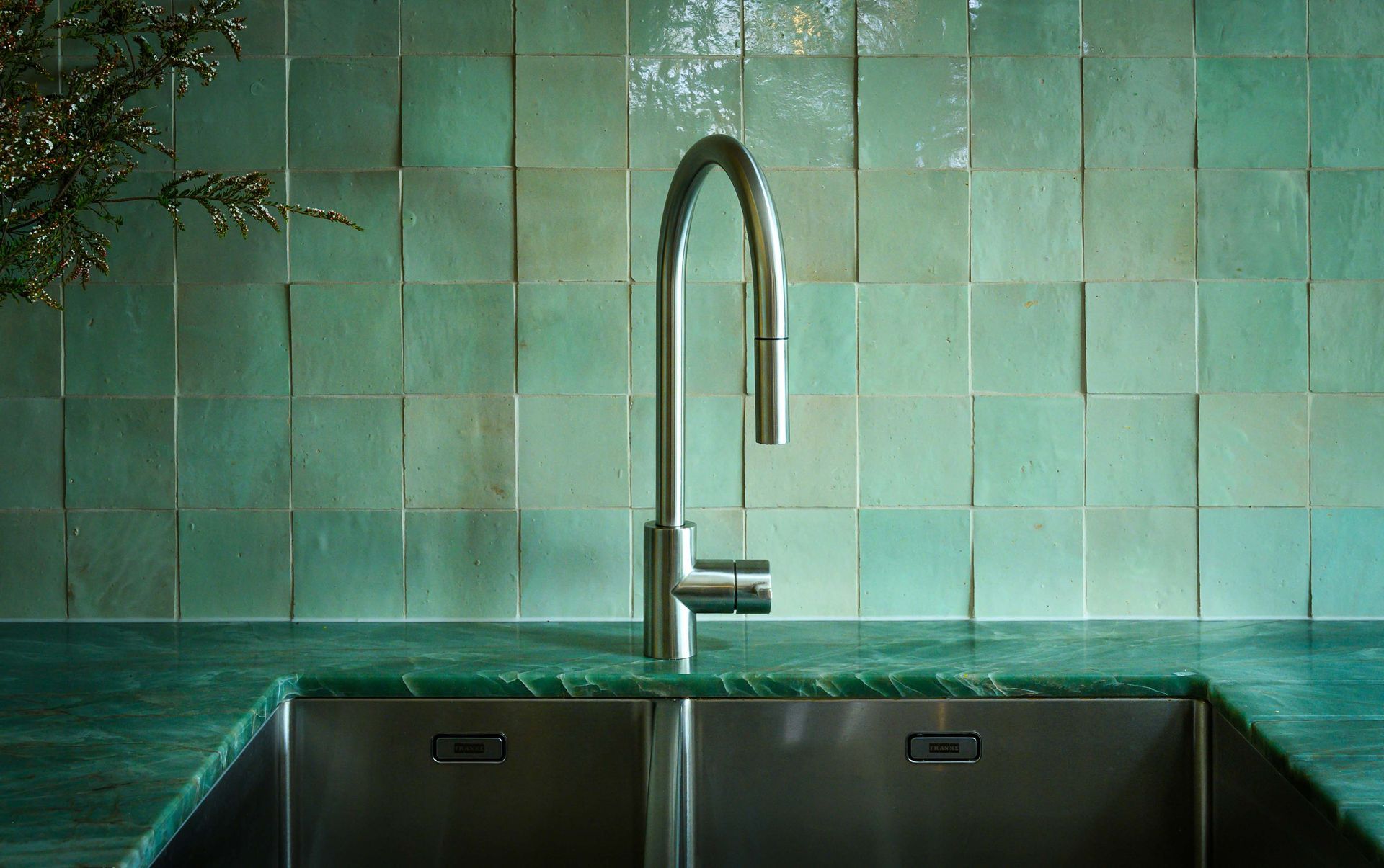 A kitchen sink with a faucet and green tiles on the wall.