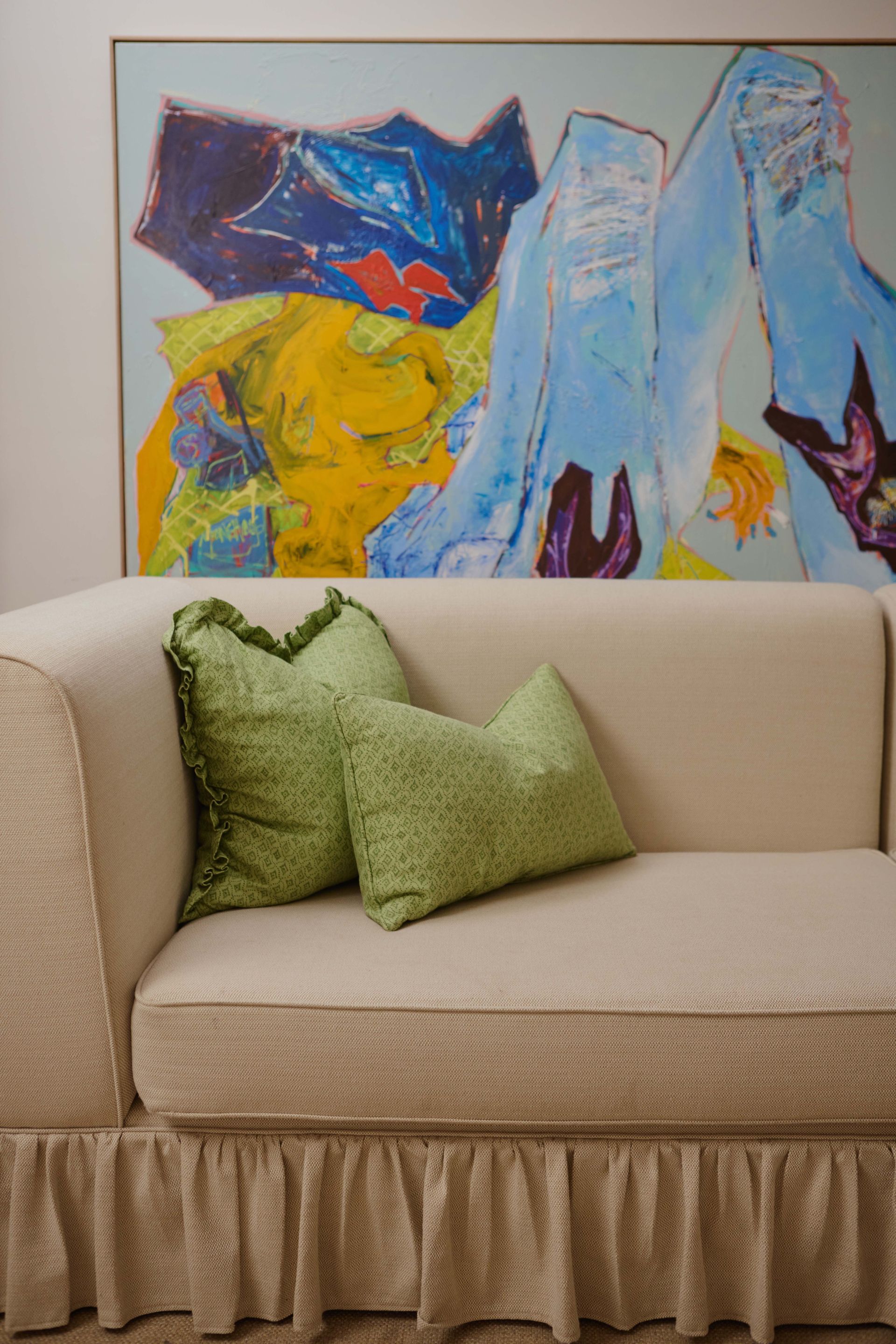 A white couch with green pillows and a painting on the wall behind it.