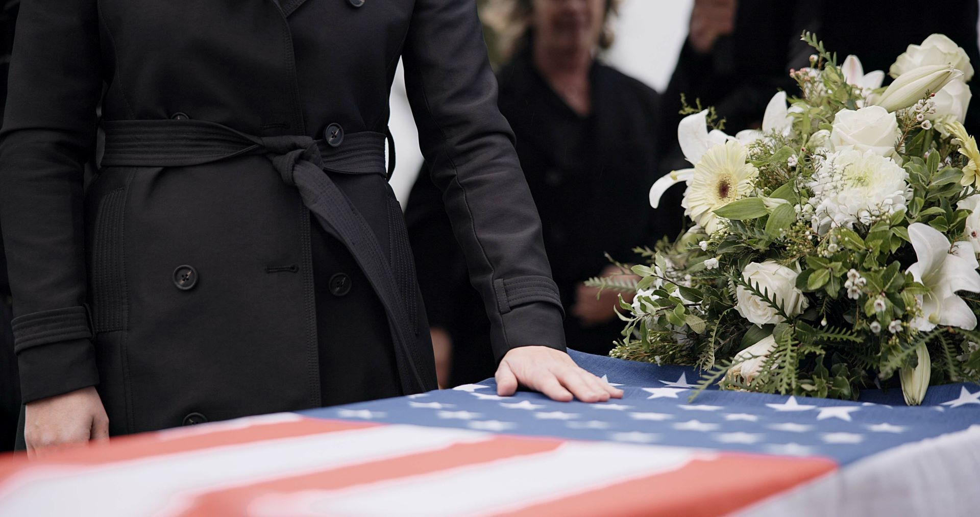 A person in a black coat rests their hand on a casket draped in an American flag, with white flowers nearby.