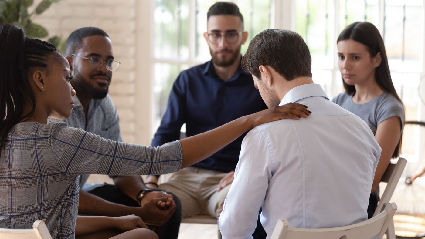A small, diverse group sits in a circle, with one person offering support by placing a hand on another’s shoulder.