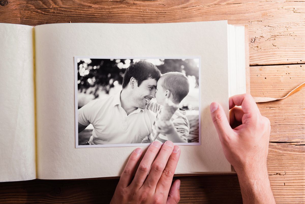 A person’s hands hold open a photo album displaying a black-and-white print of two people interacting on a wood surface.