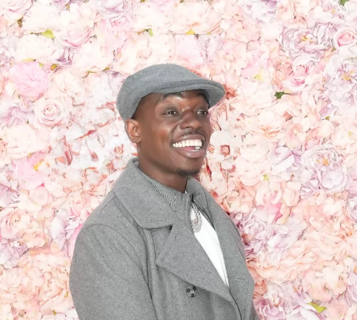 A man wearing a hat and a coat is smiling in front of a wall of pink flowers.
