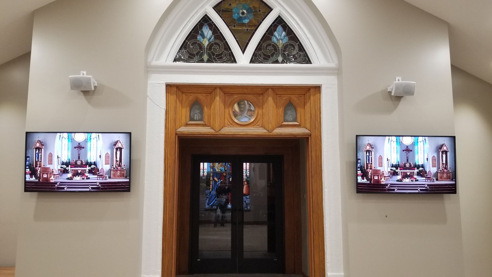 Two TVs display church service on walls flanking a wooden church door. Speakers are mounted above the TVs.