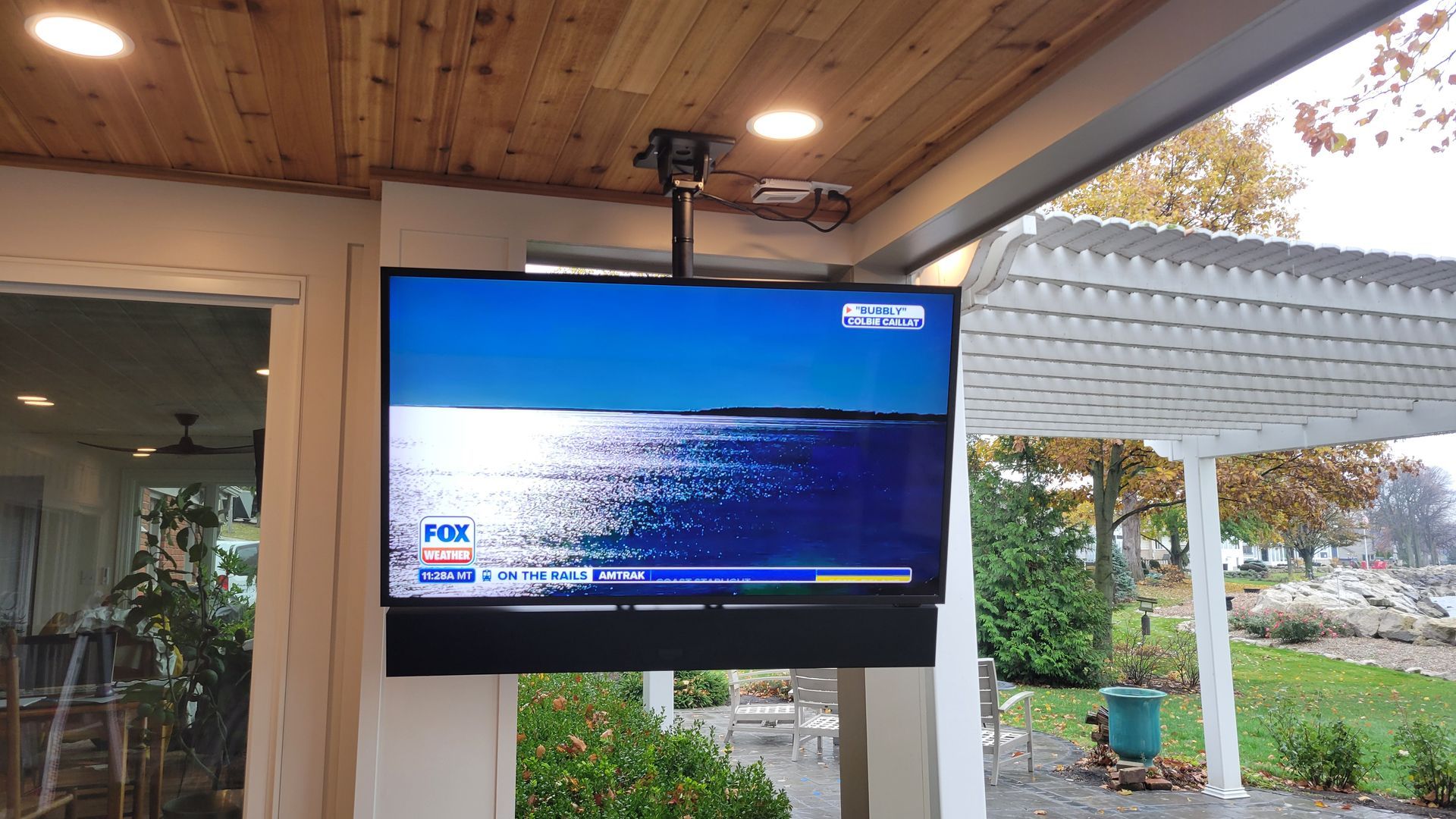 Television mounted outdoors, displaying news. Mounted on a pillar, soundbar below. Overlooking a yard and water.