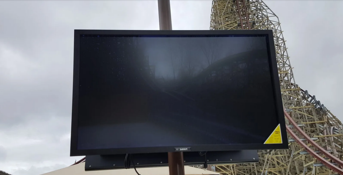 Black outdoor TV mounted outdoors, with a roller coaster in background.
