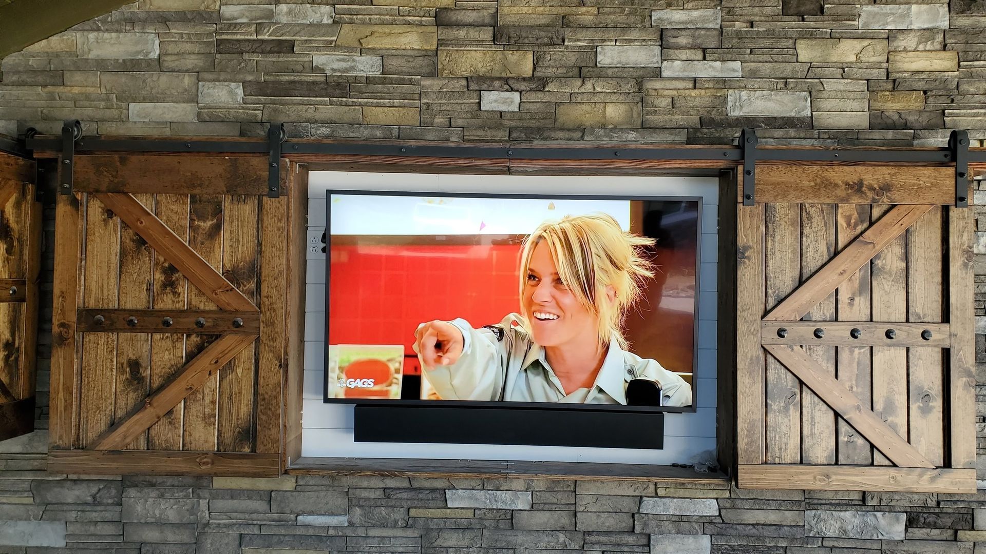 Outdoor TV custom mounted in a stone wall, framed by wooden barn doors.